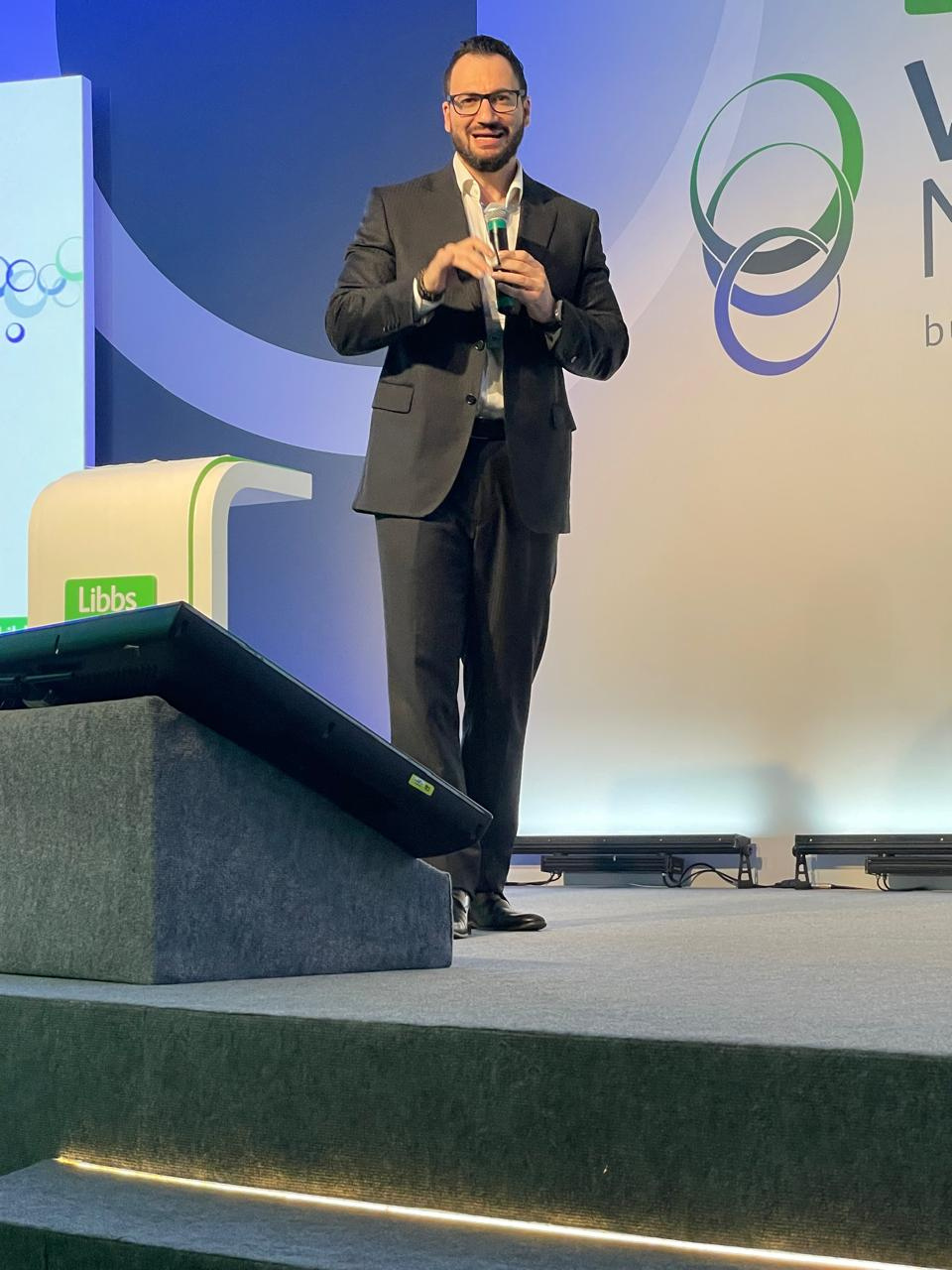 Alex Vieira de terno, no palco, conduzindo uma palestra com microfone